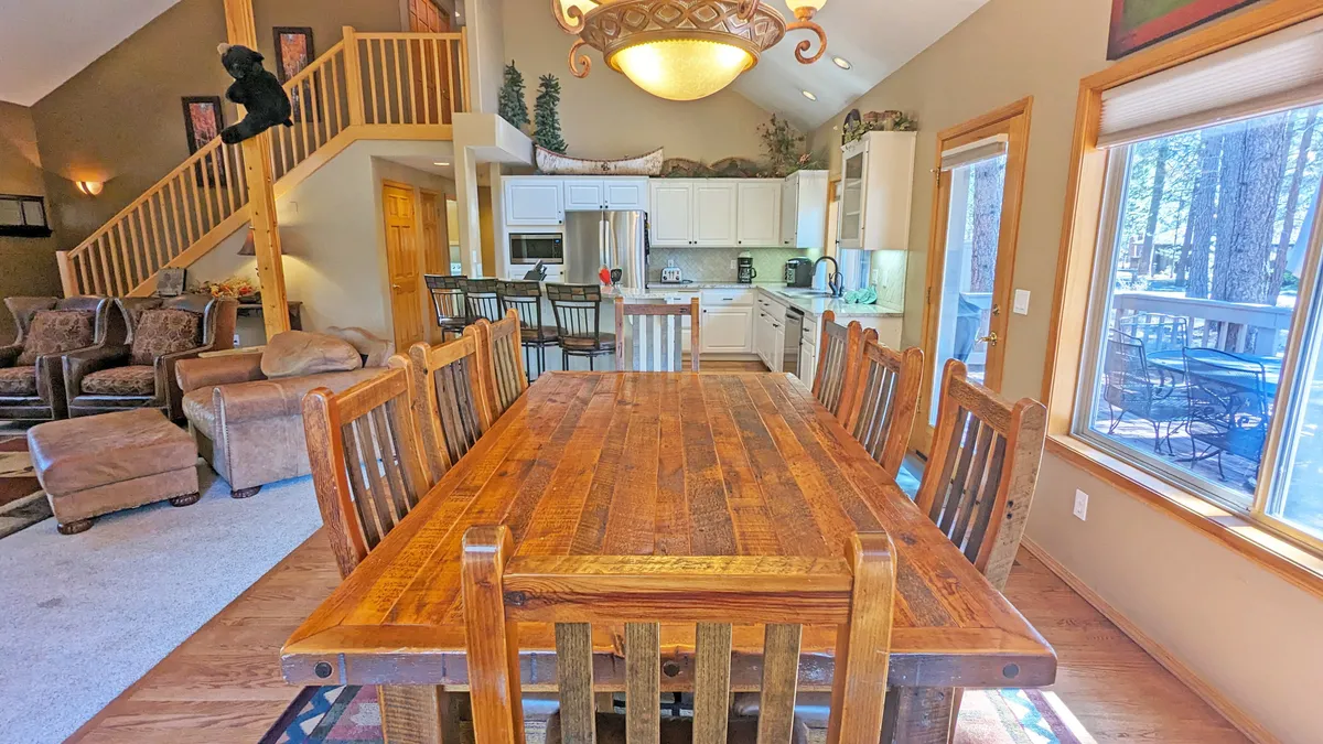 This image shows a large wooden dining table set within a spacious kitchen area, with a decorative chandelier overhead and a staircase leading to the upper level. The setting is warm and welcoming, suitable for relaxing after a day spent outdoors.
