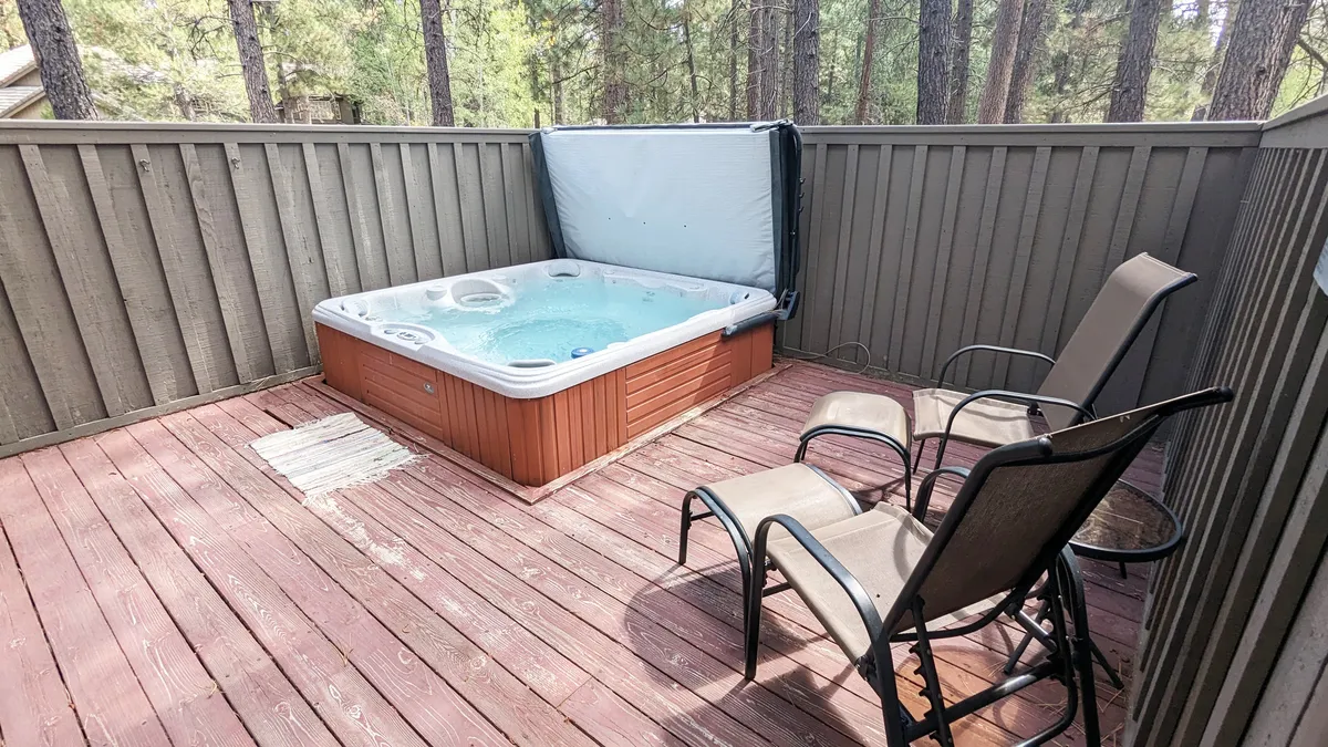An exterior shot of a bubbling hot tub on a wooden deck, surrounded by tall trees. Comfortable chairs are arranged nearby, creating a tranquil setting for relaxation or socializing under the open sky, day or night.