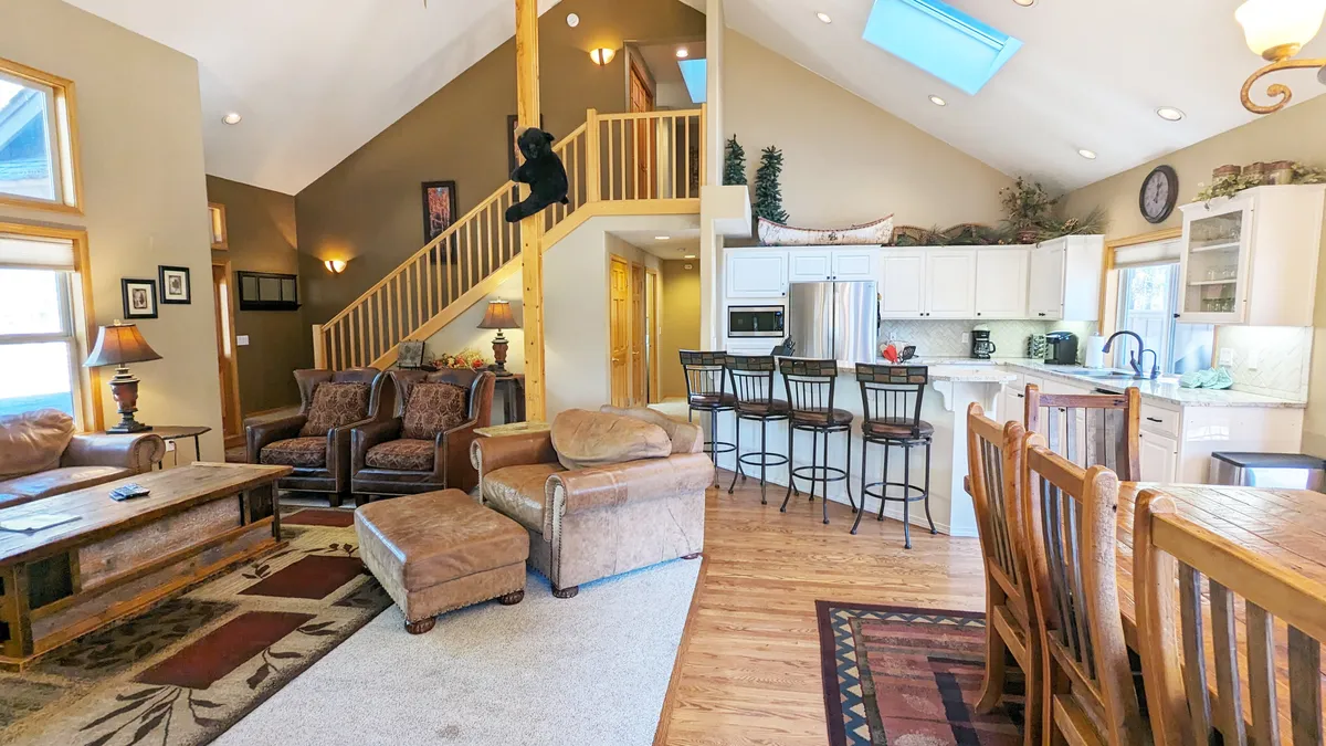A wide view of the open-plan great room showcasing a comfortable seating area with plush leather furniture and wingback chairs, an adjoining kitchen with visible new appliances and an island, a dining area with a solid wood table, and large windows letting in abundant natural light. The scene is accented by homey details like a bear decor by the staircase and a warm, inviting layout that encourages gatherings.