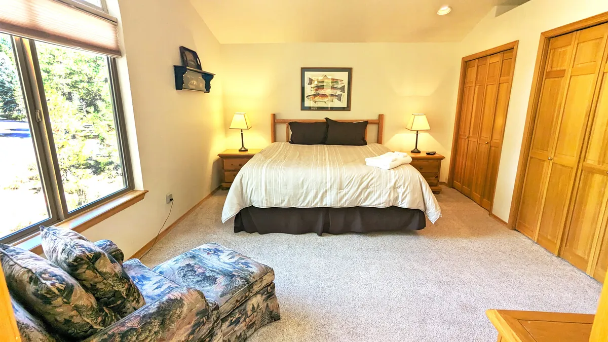 A warmly lit bedroom featuring a comfortable king-sized bed flanked by nightstands, an inviting armchair in the corner, and tasteful artwork above the headboard. Natural light streams in through a window, enhancing the cozy and restful ambiance.
