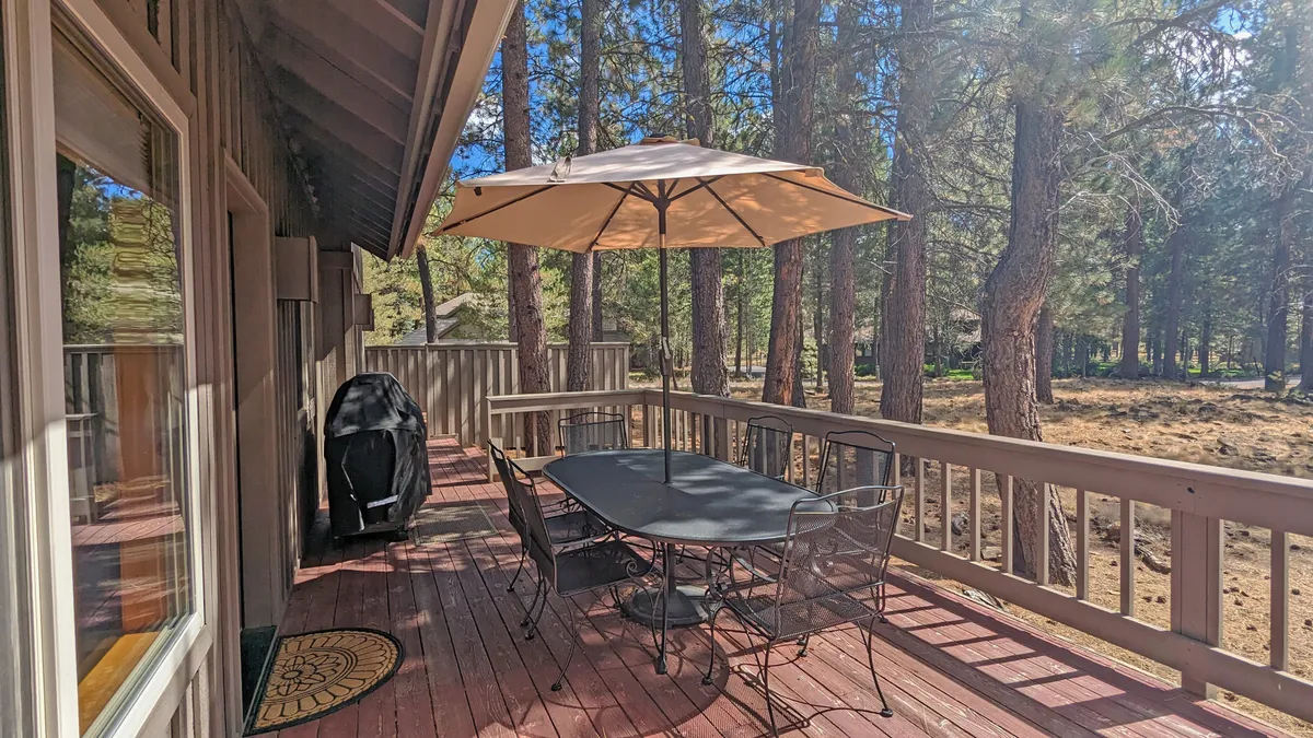 An inviting deck space equipped with a patio table and chairs, umbrella, grill, and surrounded by verdant trees. This outdoor extension connects seamlessly to the interior, highlighting opportunities for barbecuing, al fresco meals, and relaxation in nature.