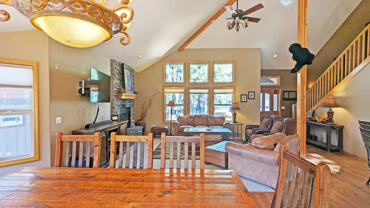 The space features large windows letting in plenty of natural light, a stone fireplace for cozy evenings, a wooden dining table, and a comfortable seating area. The design connects the indoors with the outdoors through light and views, suitable for relaxing after adventures.