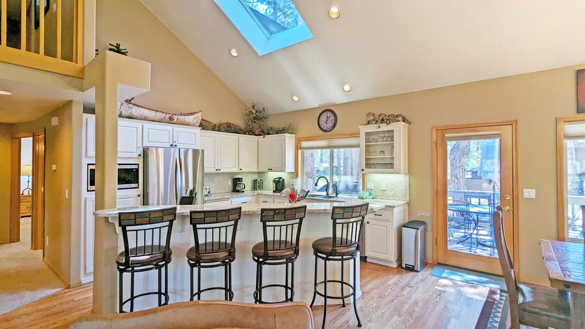 An elegant kitchen with a central island, a skylight overhead, a modern dining table, and large windows that showcase views of nature outside. The setup encourages cooking together, socializing, and sharing meals in a bright, welcoming environment.