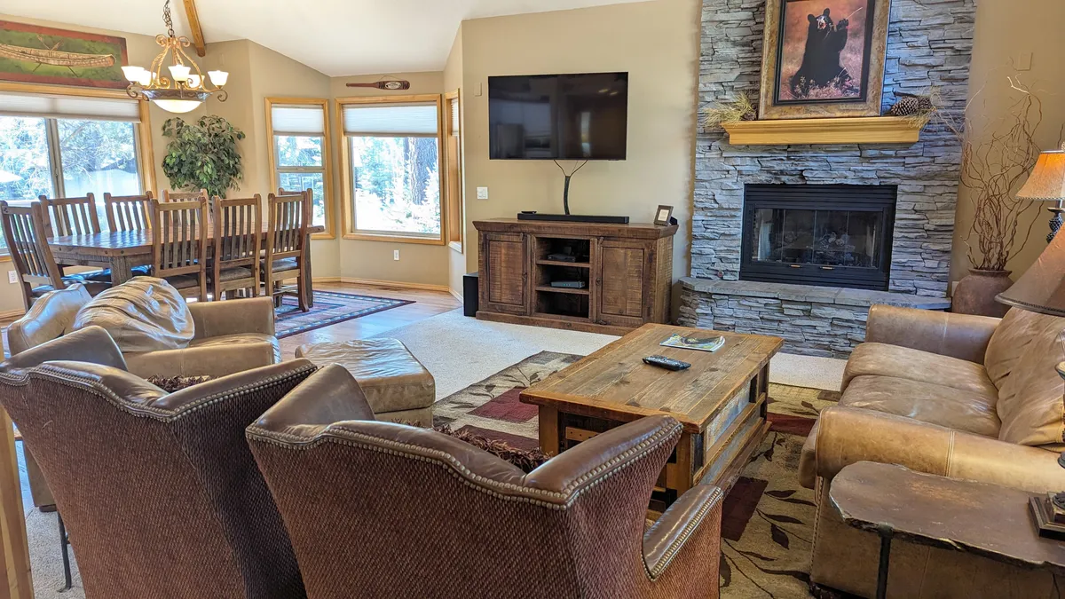 The image showcases a spacious, warmly lit living and dining area featuring a crackling fireplace, a large wooden table perfect for group meals or game nights, and a plush seating area arranged around a TV. Natural light streams in through large windows, enhancing the inviting atmosphere and offering glimpses of Sunriver’s scenic outdoors.