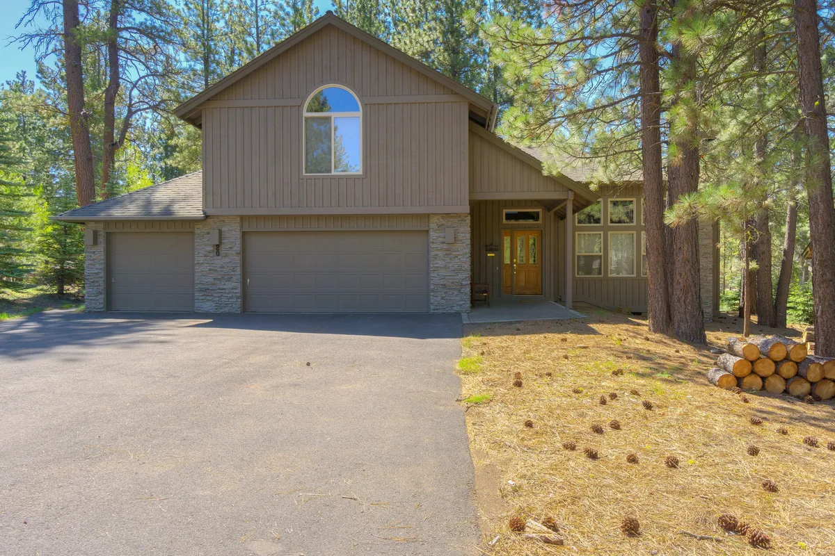 A welcoming exterior shot of the property, featuring stone accents, spacious driveway, large windows, and surrounding pine trees. The scene suggests easy access to nature and outdoor activities, emphasizing the connection between the home and the Sunriver setting.