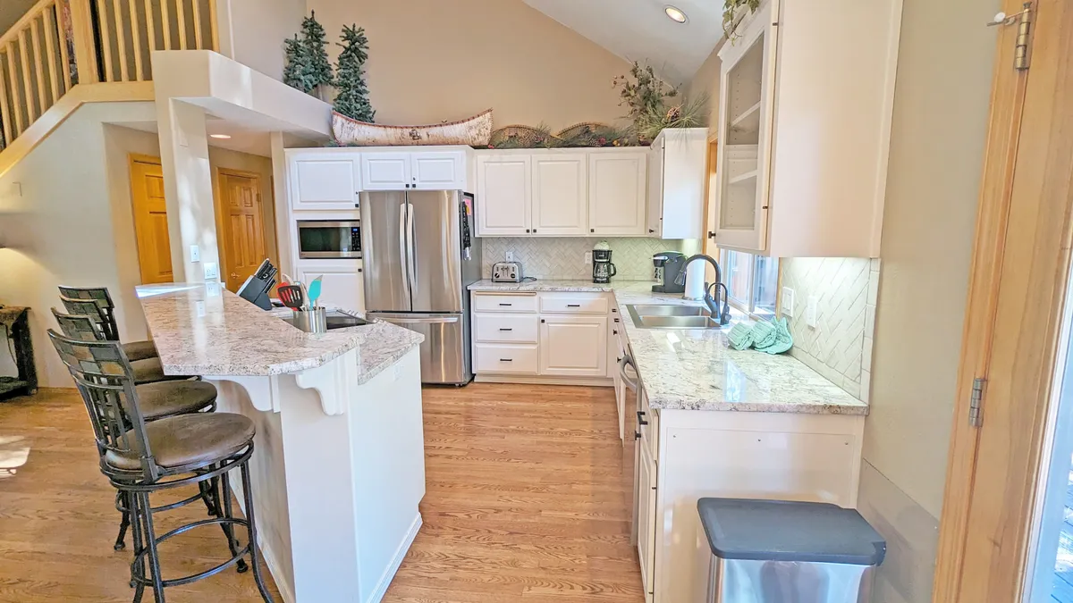 A bright, modern kitchen area with gleaming stainless steel appliances, a spacious granite countertop, kitchen island seating, and large windows emitting natural light. The scene conveys both practicality and style, with space for meal preparation and casual conversation.